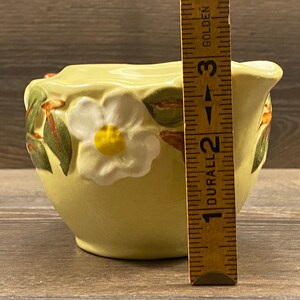 Op de afbeelding: Een gele keramische roomkan met een ontwerp van een witte bloem en groene bladeren. De roomkan is 5 cm hoog.