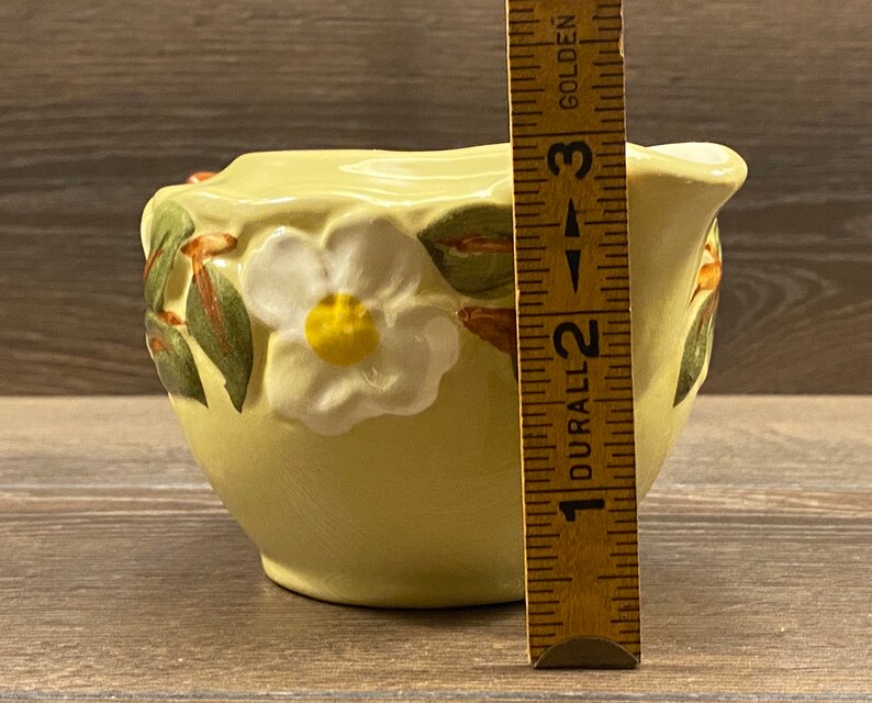 Op de afbeelding: Een gele keramische roomkan met een ontwerp van een witte bloem en groene bladeren. De roomkan is 5 cm hoog.