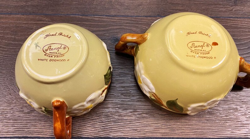 Op de afbeelding: Twee gele keramische kommen met bruine handvatten. De kommen zijn versierd met witte kornoelje bloemen en groene bladeren. De onderkant van de kommen is gemarkeerd met "Handgeschilderd" en "Stangl&reg;  Ovenvast Witte Kornoelje #".