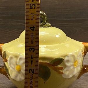 Op de afbeelding: Een gele suikerpot van keramiek met een deksel en twee handvatten. De pot is versierd met een bloemmotief met witte bloemen en groene bladeren. De pot is ongeveer 12,7 cm hoog.