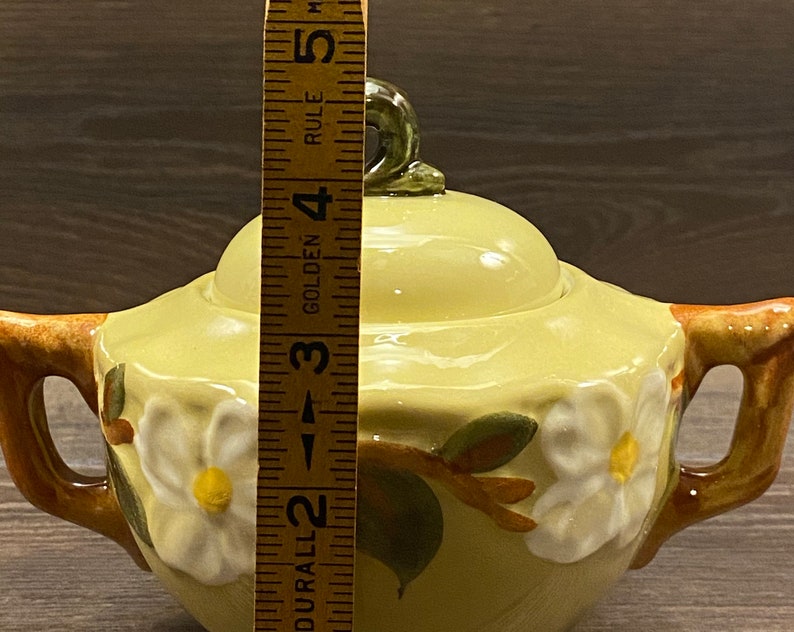 Op de afbeelding: Een gele suikerpot van keramiek met een deksel en twee handvatten. De pot is versierd met een bloemmotief met witte bloemen en groene bladeren. De pot is ongeveer 12,7 cm hoog.