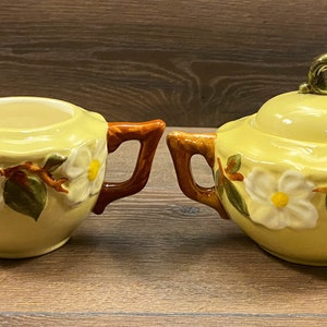 Op de afbeelding: Een vintage gele roomkan en suikerpot met een bloemenmotief. De roomkan heeft een bruine handgreep en de suikerpot heeft een gouden knop op de deksel.