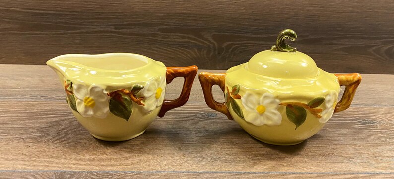 Op de afbeelding: Een vintage gele roomkan en suikerpot met een bloemenmotief. De roomkan heeft een bruine handgreep en de suikerpot heeft een gouden knop op de deksel.