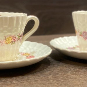 May include: Two white teacups and saucers with a floral pattern. The cups have a scalloped rim and a woven design. The saucers have a scalloped edge.