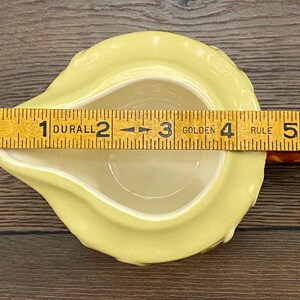 Op de afbeelding: Een gele keramische roomkan in de vorm van een peer met een handvat. De roomkan is 7,6 cm breed en 10,2 cm lang. De roomkan staat op een houten oppervlak.
