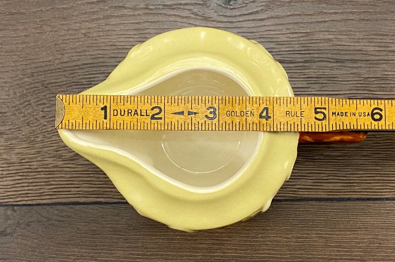 Op de afbeelding: Een gele keramische roomkan in de vorm van een peer met een handvat. De roomkan is 7,6 cm breed en 10,2 cm lang. De roomkan staat op een houten oppervlak.
