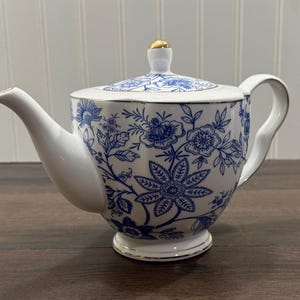 May include: White ceramic teapot with a blue floral pattern. The teapot has a curved spout, a handle, and a lid with a gold knob. The base and rim are trimmed with gold. The design features detailed blue flowers and foliage.