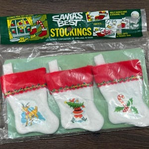 May include: Three small Christmas stockings in a plastic package. The stockings are white with red tops and feature festive designs of a reindeer, Santa Claus, and a candy cane. The package reads "Santa's Best Stockings."