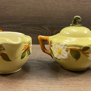 Op de afbeelding: Een vintage gele room- en suikerset met een bloemenontwerp. De set heeft witte bloemen met groene bladeren en bruine stelen. De roomkan heeft een gebogen handvat en de suikerpot heeft een deksel met een groene knop.