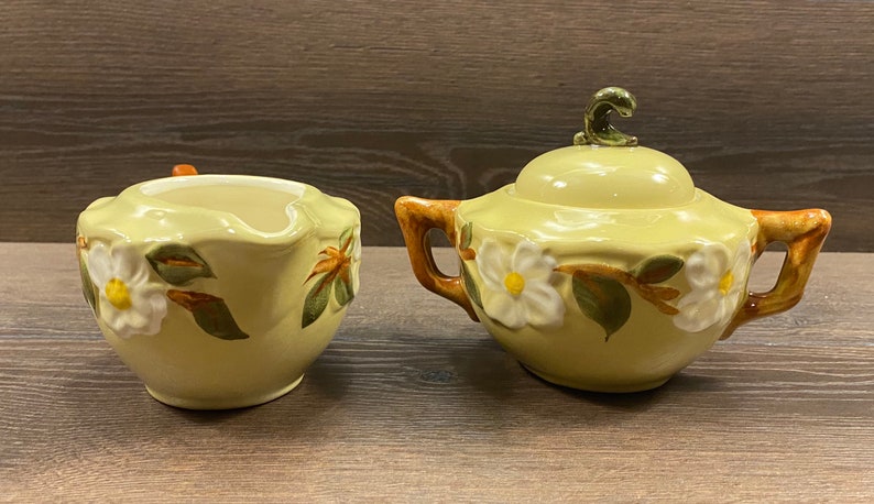 Op de afbeelding: Een vintage gele room- en suikerset met een bloemenontwerp. De set heeft witte bloemen met groene bladeren en bruine stelen. De roomkan heeft een gebogen handvat en de suikerpot heeft een deksel met een groene knop.