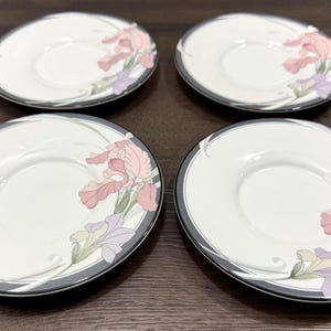 May include: Four white ceramic saucers with a gray border and floral iris design in pink, purple, and green. The saucers are arranged on a dark brown surface.