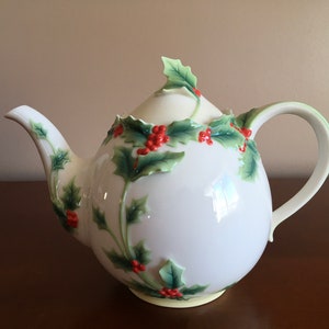 May include: White porcelain teapot featuring a detailed design of holly leaves and red berries. The teapot has a round body and a curved handle.