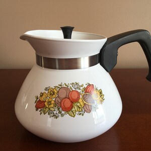 May include: White enamel teapot with a black handle and lid knob. The teapot features a floral design with orange, yellow, and brown flowers and fruit.
