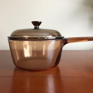 May include: A brown glass saucepan with a lid and a handle. The saucepan is on a wooden surface.