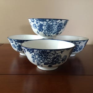 以下が含まれることがあります： 青い花柄の白い陶器のボウルが4つ。ボウルは濃い茶色の木製の表面に配置されています。花柄は、花と葉の精巧な描写です。
