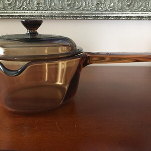 May include: A translucent amber-coloured glass saucepan with a matching lid. The pot features a long, integrated glass handle and a small pouring spout on the side. The glass is smooth and tinted in a warm, brownish-orange hue throughout.
