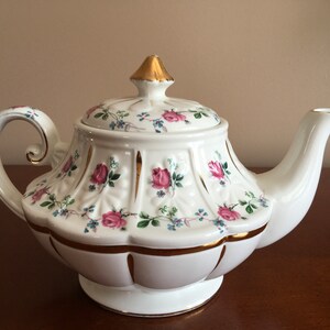May include: White teapot with a gold trim and floral pattern featuring pink roses and green leaves. The teapot has a lid with a gold finial.