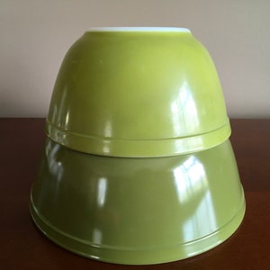 May include: Two green glass mixing bowls, one nested inside the other. The bowls are a muted shade of green with a glossy finish.