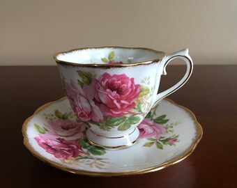 May include: A white teacup and saucer with a pink rose floral pattern and gold trim.