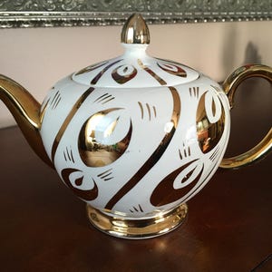 May include: A white ceramic teapot featuring a reflective gold metallic pattern of stylised leaf or petal shapes. The teapot includes a matching lid with a gold finial, a gold-trimmed spout, a gold handle, and a gold base rim.