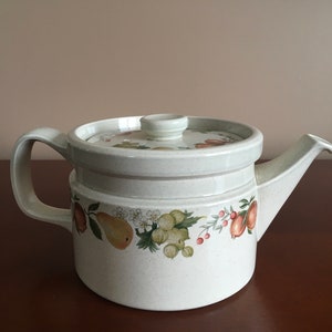 Peut inclure: Une théière en céramique blanche avec un couvercle et un motif floral représentant des poires, des pommes et des baies. La théière a une poignée et un bec.