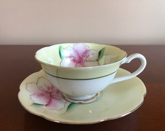 以下が含まれることがあります： ピンクの花柄の黄色いティーカップとソーサーのセット。カップは白い内側と金の縁取りがあります。ソーサーは波打った縁取りがあります。