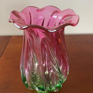 May include: A decorative glass vase with a ruffled rim. The vase is a vibrant pink colour that fades into green at the base. The glass has a swirled design, adding texture and visual interest to the piece.