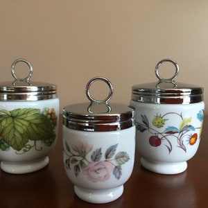 May include: Three white ceramic egg coddlers with silver lids and floral designs. The coddlers have a ring handle on the lid. The designs include a green grapevine, a pink rose, and red strawberries.