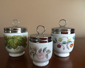 Pode incluir: Três coqueteleiras de cerâmica branca com tampas prateadas e desenhos florais. As coqueteleiras têm uma alça de anel na tampa. Os desenhos incluem uma videira verde, uma rosa rosa e morangos vermelhos.