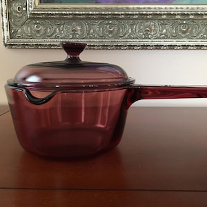 May include: A translucent deep purple glass saucepan with a matching lid and a long, integrated handle. The circular pot features a small pouring spout on the side, and the lid is topped with a small, rounded knob for easy lifting and handling.