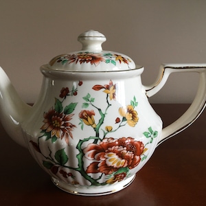 May include: A white teapot with a floral design in shades of brown, yellow, and green. The teapot has a gold trim around the lid and base.