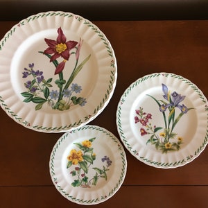 May include: Three white ceramic plates with green trim and floral designs. The largest plate features a red flower with yellow centre, the middle plate features a purple flower, and the smallest plate features a yellow flower.