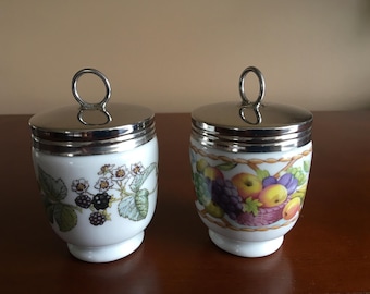 Pode incluir: Dois coqueteleiras de cerâmica branca com tampas prateadas. Uma coqueteleira apresenta um desenho floral com amoras, enquanto a outra tem um desenho de frutas com maçãs, uvas e ameixas.