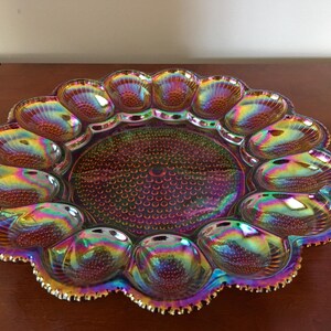 May include: A round iridescent carnival glass egg plate featuring a central circular section with a hobnail pattern surrounded by twelve individual egg-shaped indentations, each with a ribbed design and a scalloped, serrated outer edge.
