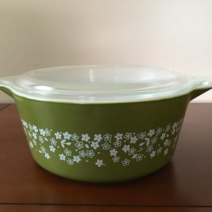 May include: A green Pyrex bowl with a clear glass lid. The bowl features white floral patterns around the outside. It rests on a dark brown wooden surface.