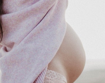 May include: A pregnant woman wearing a light pink sweater and white lace underwear stands on a sandy beach. She is looking off to the side and has her hand on her head.