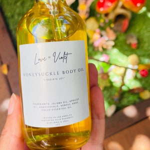 May include: A clear glass bottle of honeysuckle body oil with a black dropper. The label reads "Love & Violet" and "Honeysuckle Body Oil". The oil is yellow, and the ingredients include jojoba oil and violet essential oil. The bottle is 4 oz.