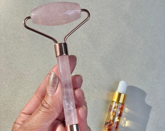 May include: A rose quartz facial roller with a rose gold handle. The roller is held in a hand.