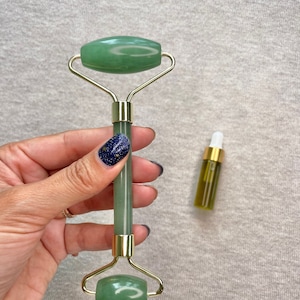 May include: A green jade roller with a gold handle. The roller is held in a hand with green nail polish. A small bottle of oil is in the background.