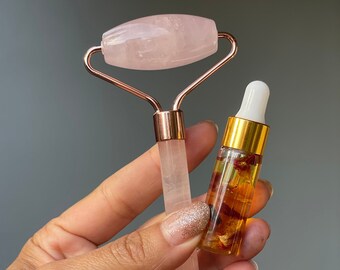 May include: A rose quartz facial roller with a gold-toned handle and a small bottle of rose oil with dried rose petals inside. The roller is held in a hand.