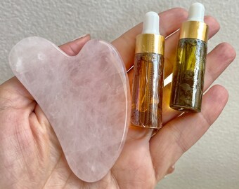 May include: A pink rose quartz gua sha tool and two amber glass bottles with white droppers. The bottles and tool are held in a hand.