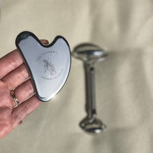 以下が含まれることがあります： 「The Alchemy Goddess Collective」と刻まれた銀色のハート型の顔用gua shaツールです。