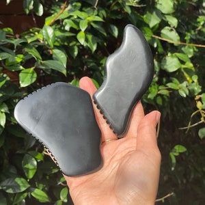 May include: Two black gua sha tools, one shaped like a heart and the other shaped like a fish, are held in a person's hand. The tools are made of smooth, polished black stone.