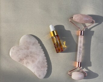 May include: A rose quartz Gua Sha tool, face roller, and small glass bottle with a dropper. The Gua Sha is heart-shaped. The face roller has a rose gold handle. The bottle contains a clear liquid with visible inclusions.