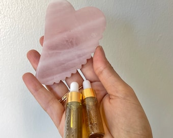 May include: Two small amber glass bottles with gold caps and droppers, containing a clear liquid, are held in a hand. A pink heart-shaped stone is also in the hand.
