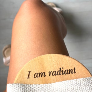 Puede incluir: Un cepillo corporal de madera con una correa blanca. El cepillo tiene las palabras "Soy radiante" grabadas.