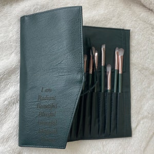 May include: A dark green leather makeup brush roll with a zipper closure. The roll has multiple slots for brushes and has the text "I am Radiant Beautiful Blessed & Worthy" engraved on the front.