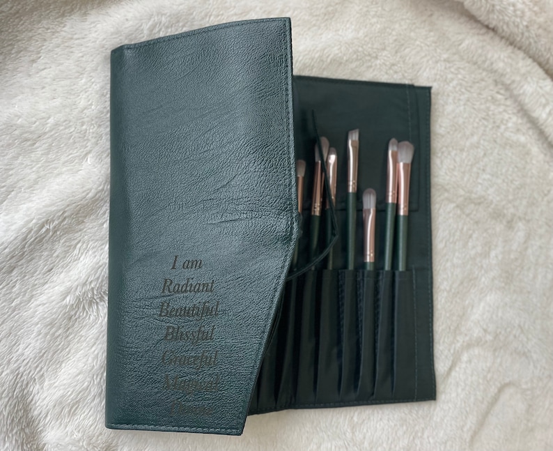 May include: A dark green leather makeup brush roll with a zipper closure. The roll has multiple slots for brushes and has the text "I am Radiant Beautiful Blessed & Worthy" engraved on the front.