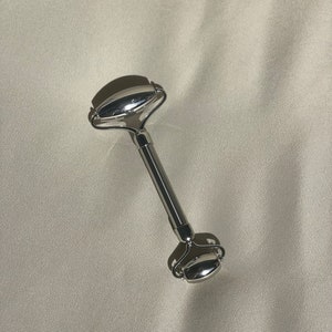 May include: A silver facial roller with two smooth, oval-shaped rollers. The roller is designed to be used on the face and neck to help reduce puffiness and improve circulation.