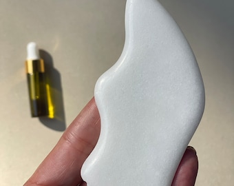 May include: White jade gua sha tool with a smooth, rounded surface and a textured edge. The tool is shaped like a face and is held in a hand.